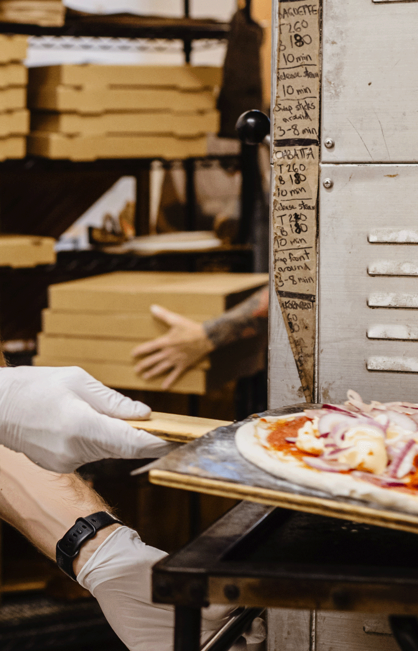 busy pizza kitchen with staff member loading pizza into cardboard box while wearing white disposable glvoes.