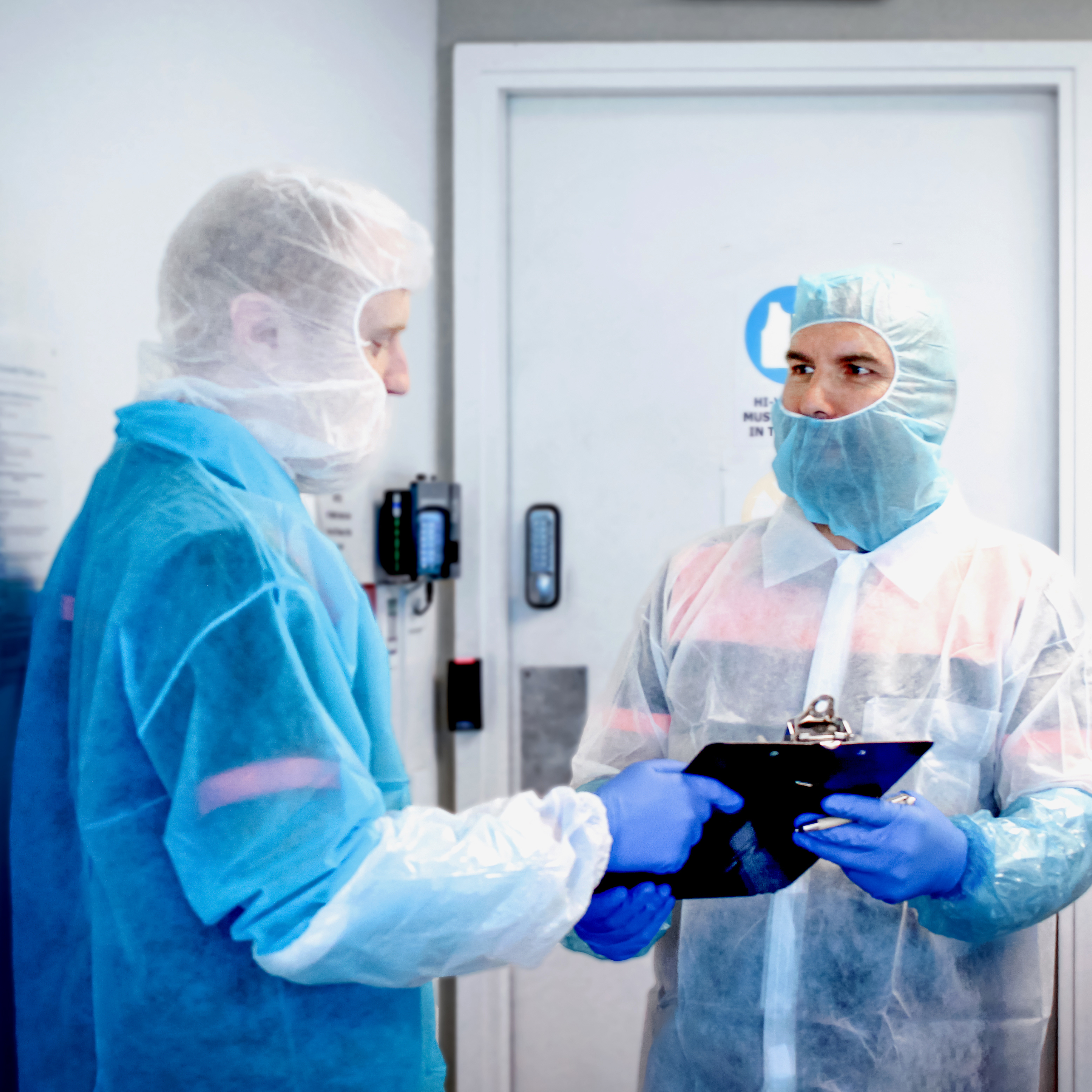 two male staff members in hallway discussing over clipboard wearing balaclavas, lab coats, sleeve covers and nitrile gloves (all disposable)