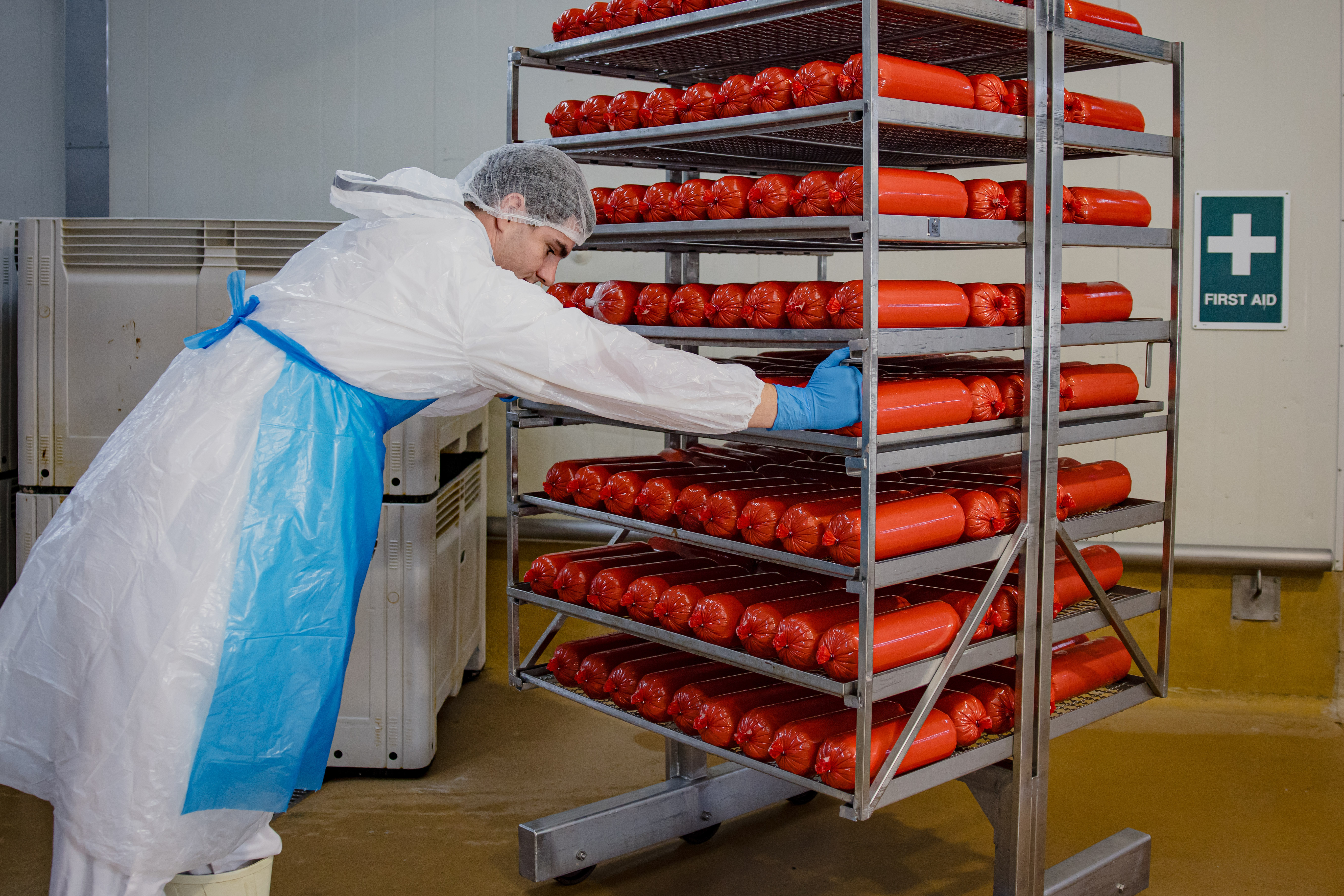 A meat worker wearing a Bastion splash jacket, apron, long cuff gloves and a clip hat, pushing a tray of meat to the ovens