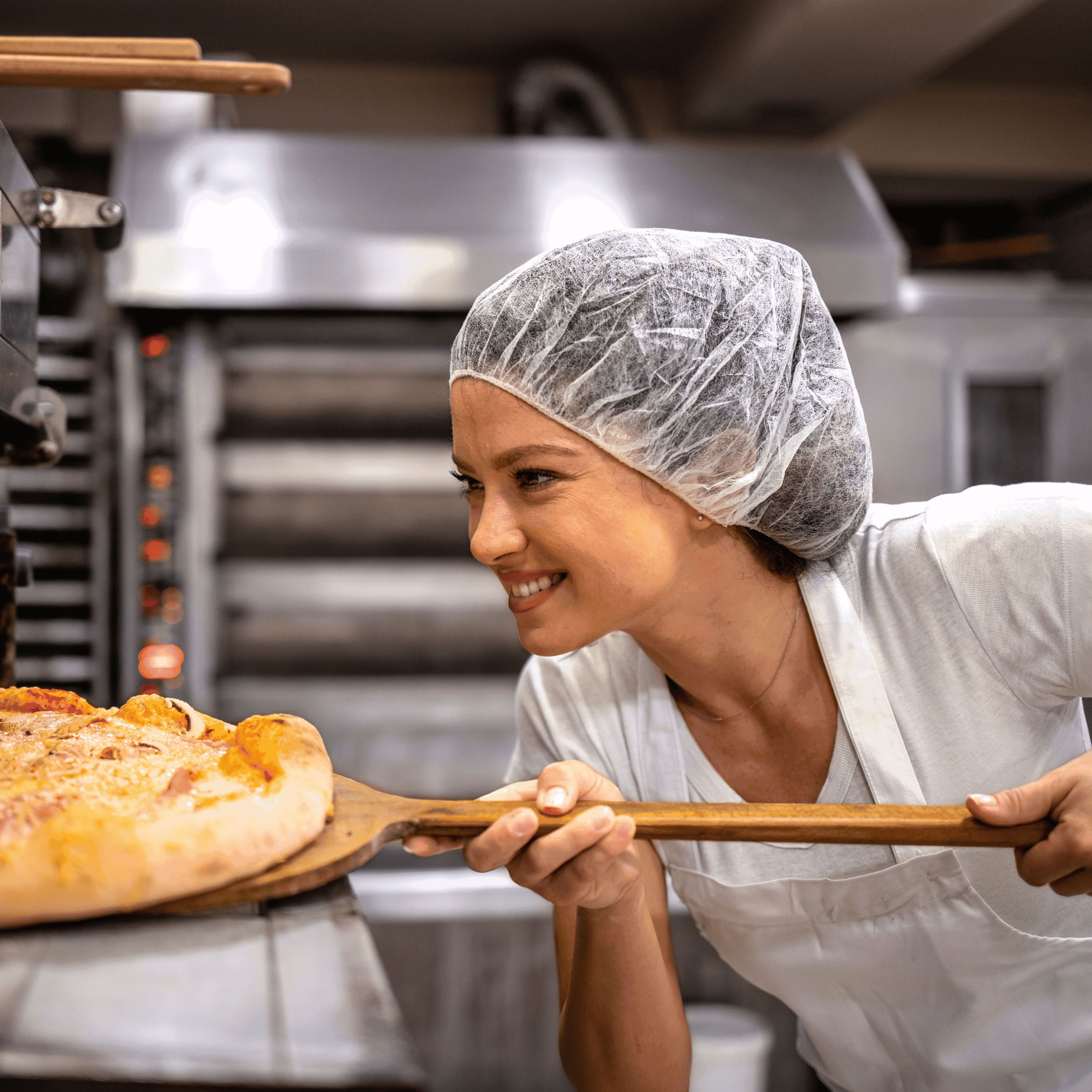 smiling female wearing bouffant hat removing pizza from oven