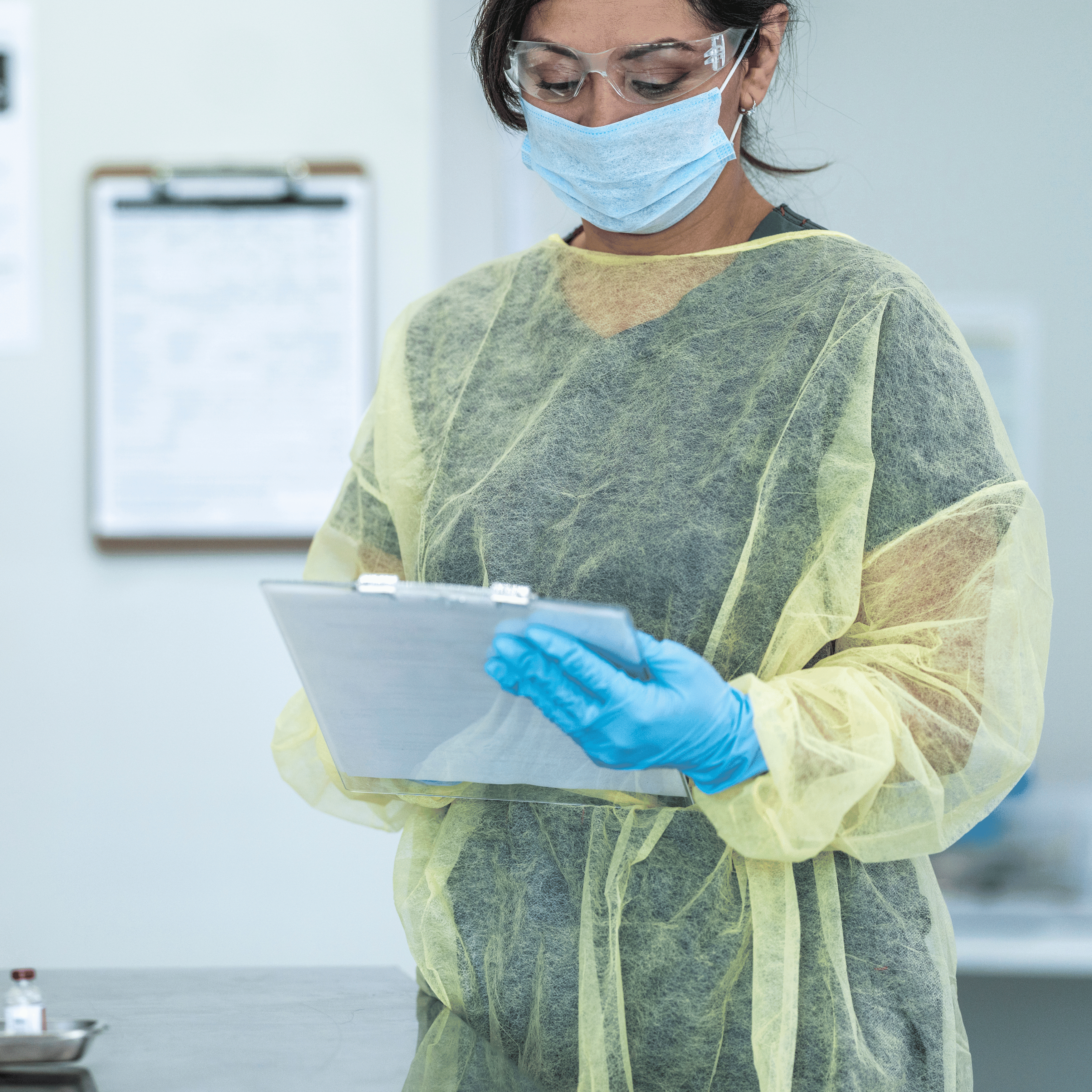 Medical staff member holding clipboard wearing blue disposable gloves, yellow isolation gown, mask and goggles.