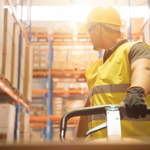 A warehouse worker wearing a hard hat and a hi-vis vest, pulling a hand trolley