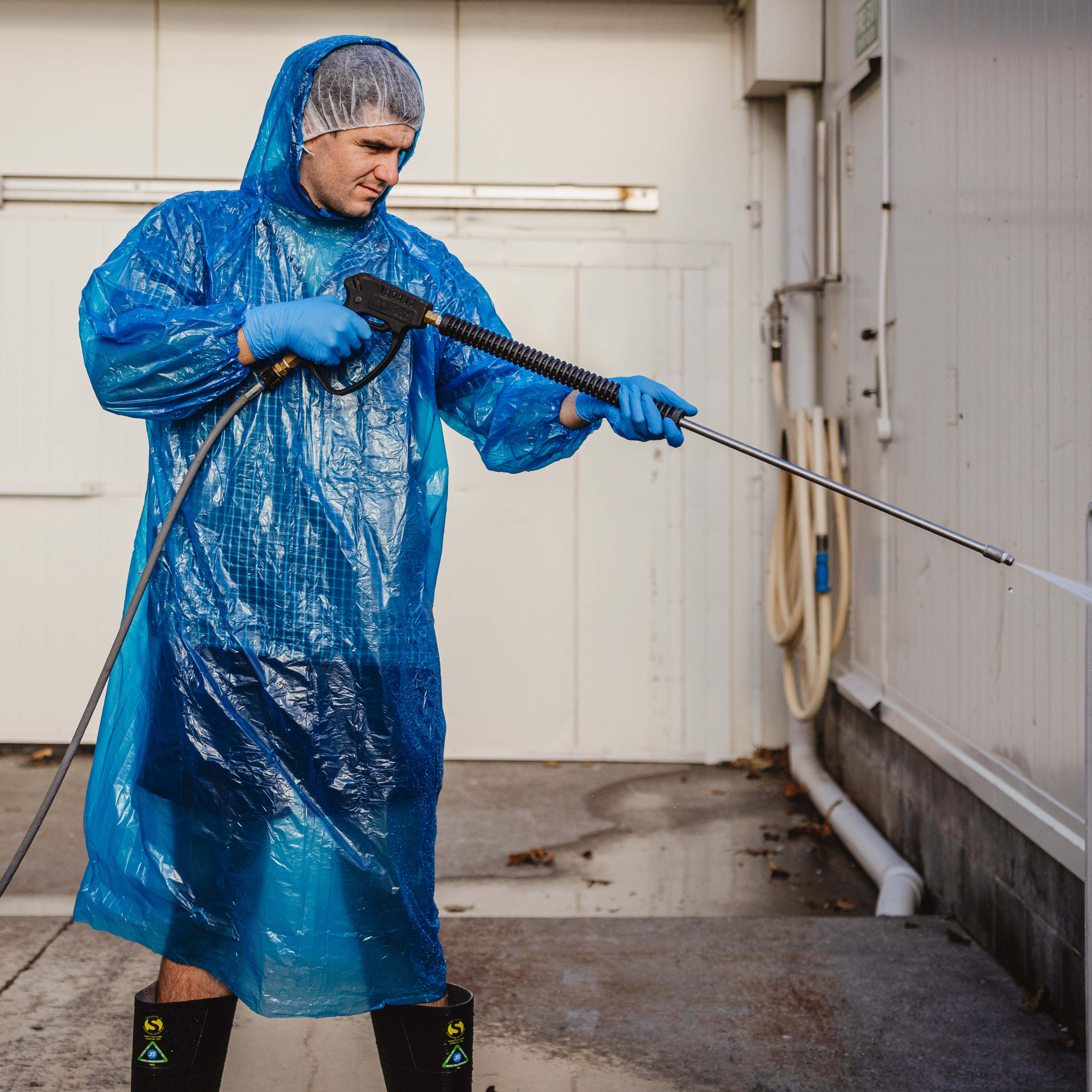 man wearing blue longsleeve splash jacket, nitrile gloves and hairnet and black gum boots while pressure washing something outdoors.