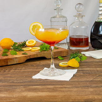 Orange and red Cocktail in a martini glass, garnished with lemon, sitting on a white Green Choice cocktail napkin. Surrounded by a chopping board with chopped Lemons and mint leaves, decanters in the background.