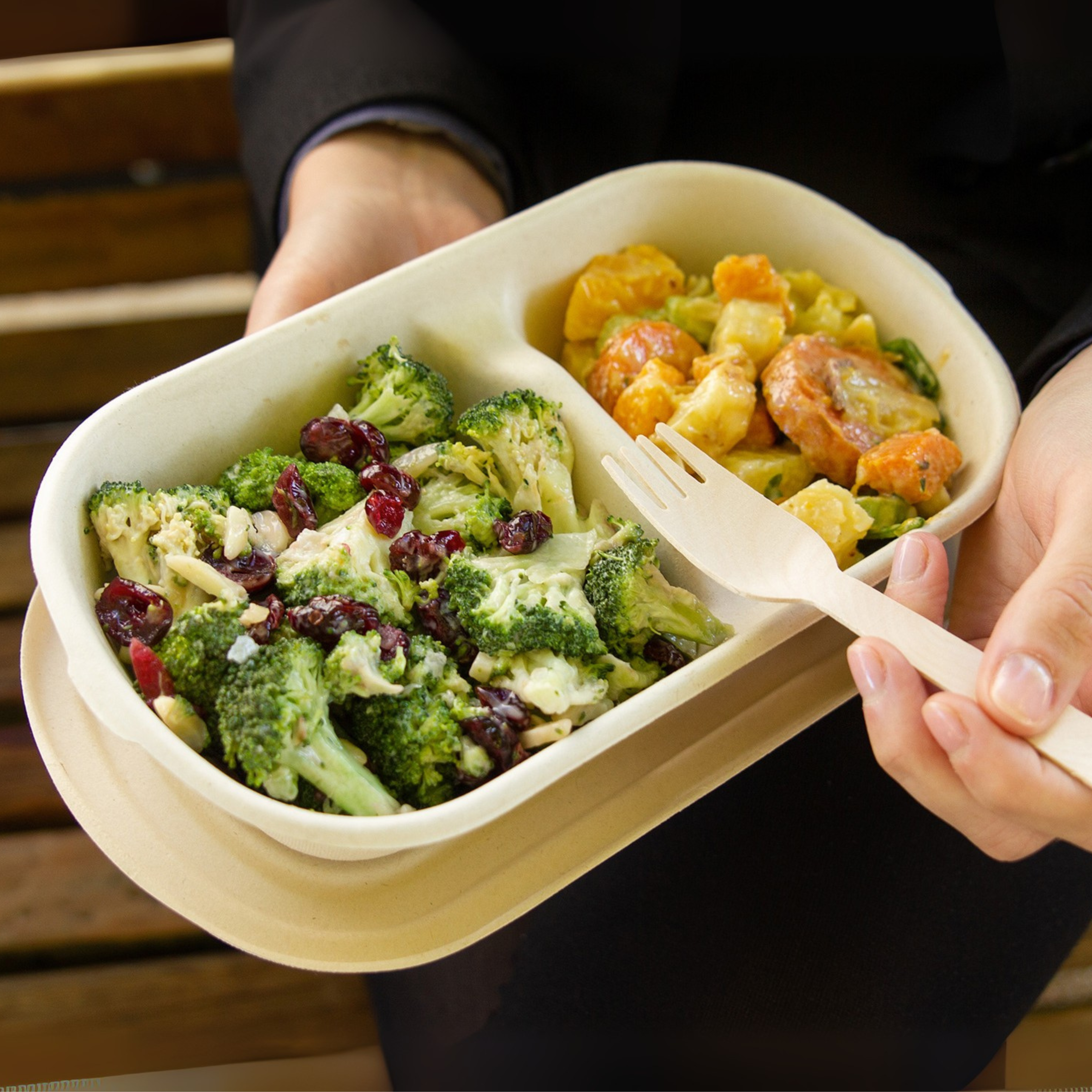 A close-up of someone sitting on a park bench eating from a Green Choice 2 compartment Food Box