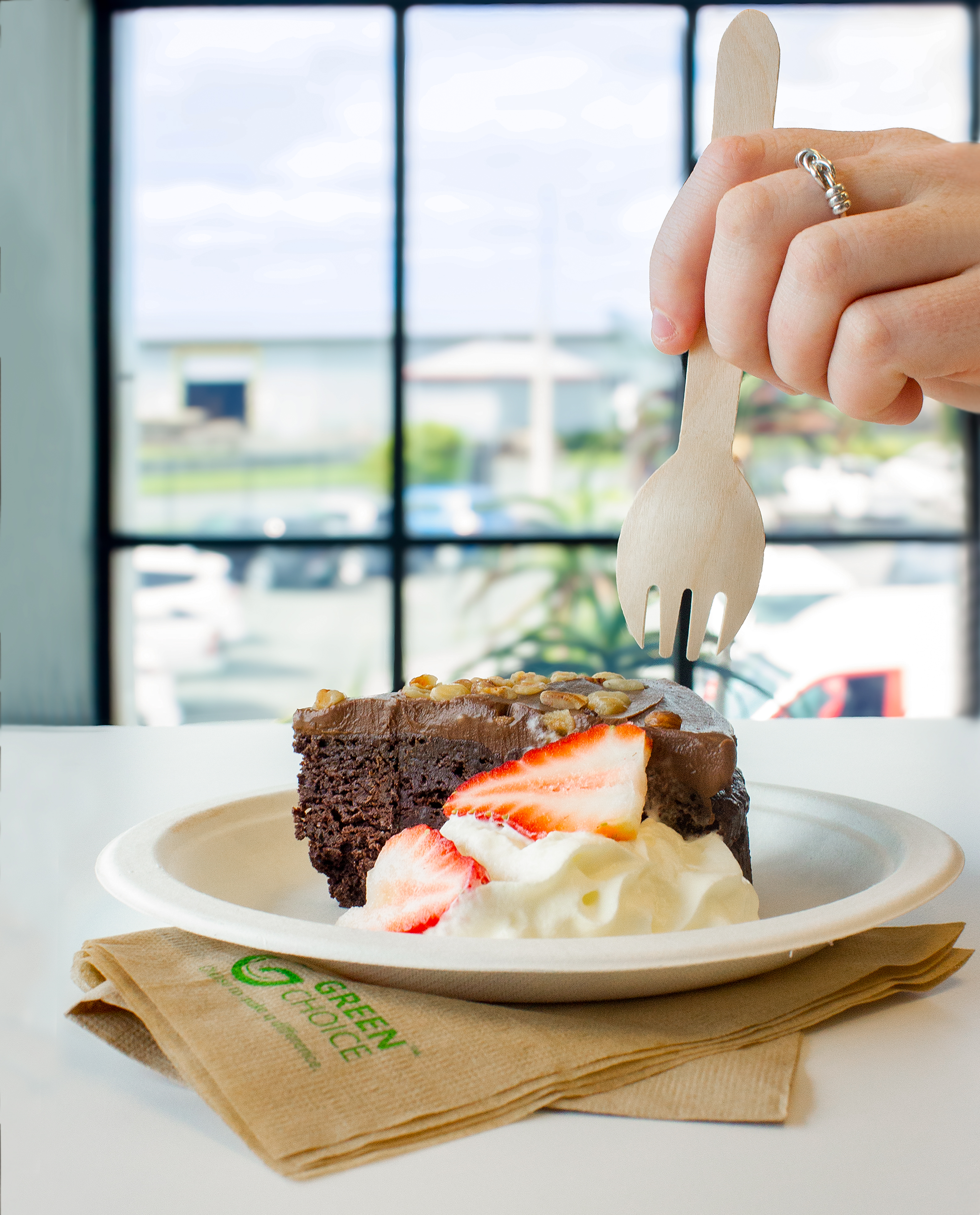 Close-up of a slice of cake with some whipped cream and strawberries sitting in a Green Choice Sugarcane plate, a hand holding a Green Choice Spork is ready to take a spoonful