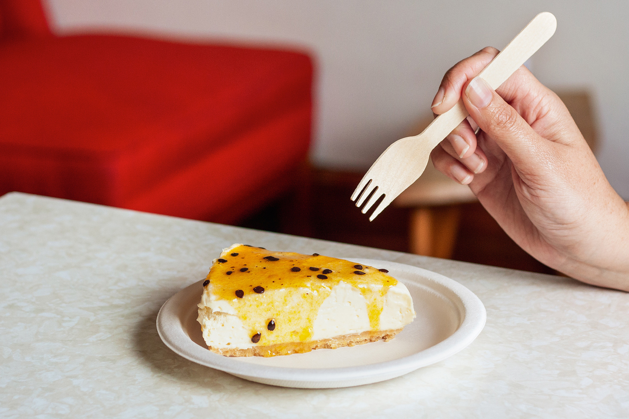 Someone holding an Unbranded Green Choice Wooden Fork, ready to take a forkful of cheesecake that is sitting in a Green Choice Sugarcane plate.