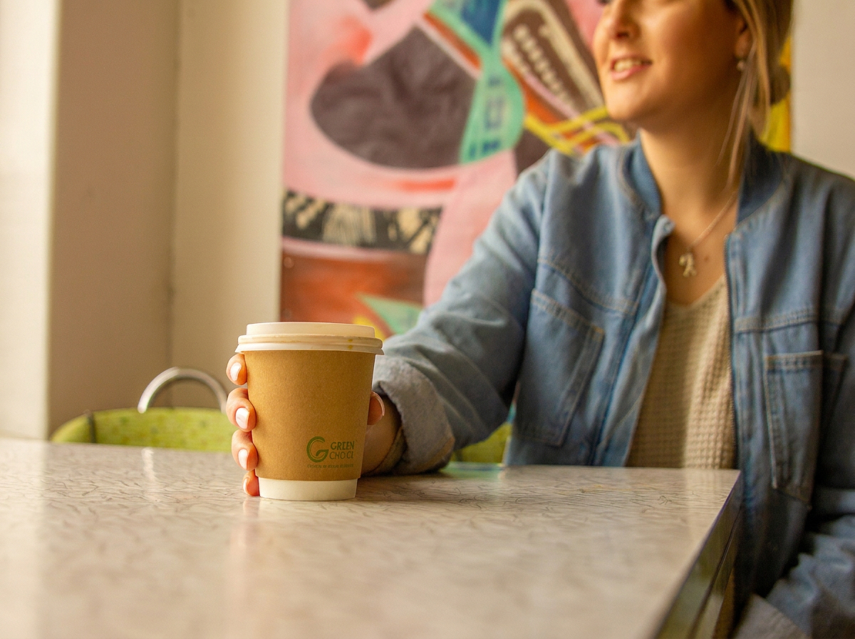 A lady sitting in a cafe holding a Green Choice Coffee Cup, looking out a window