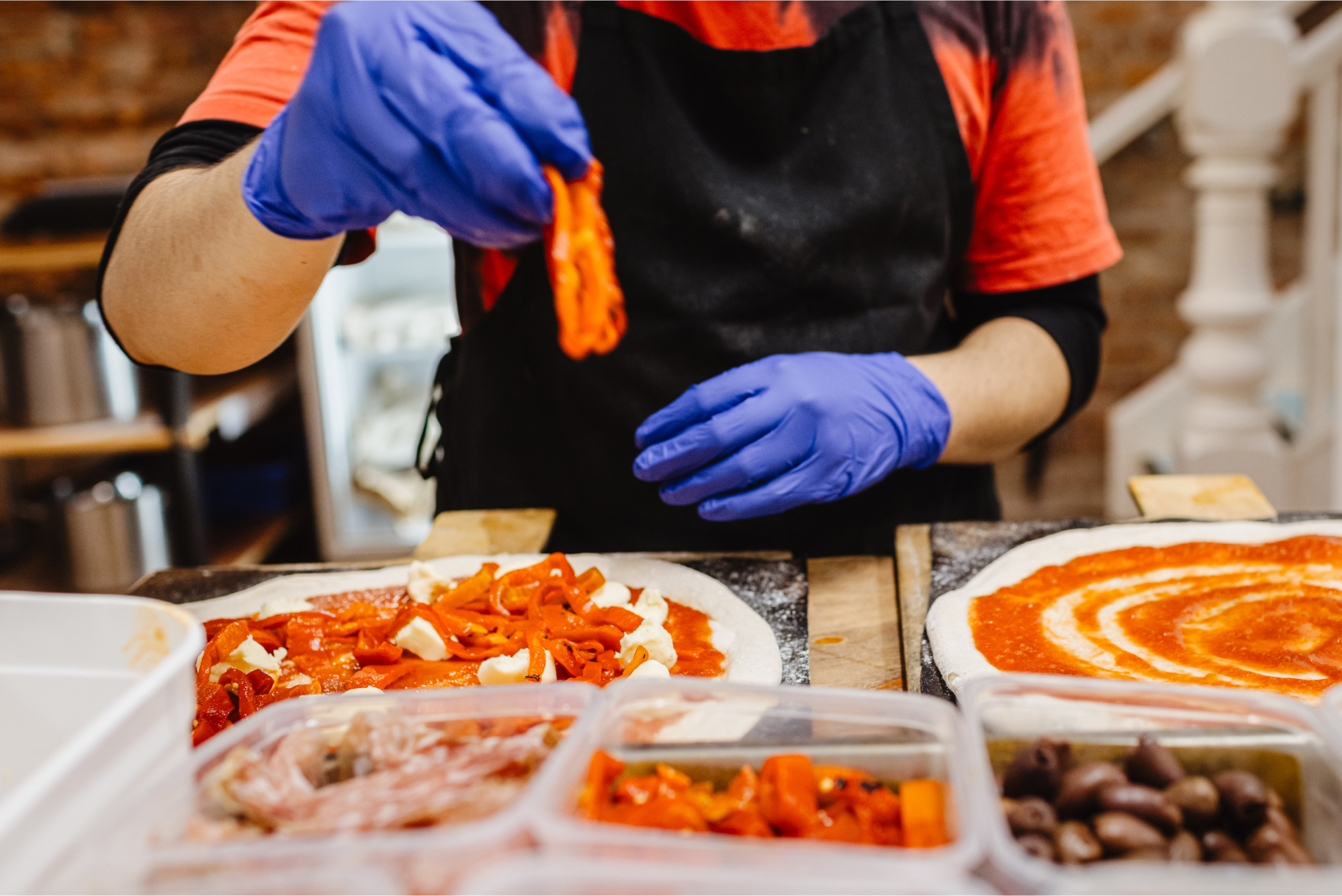 nitrile gloved hands assembling pizzas with clear containers of ingredients in view at prep station.
